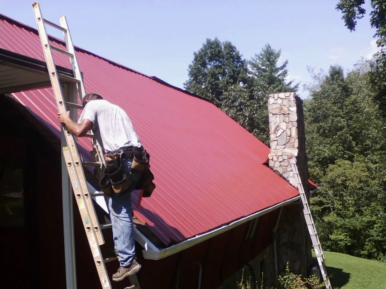 Roofer on a ladder beside a red metal roof and stone chimney — Soffit & Fascia Repair by Roofing Brothers in Tivoli