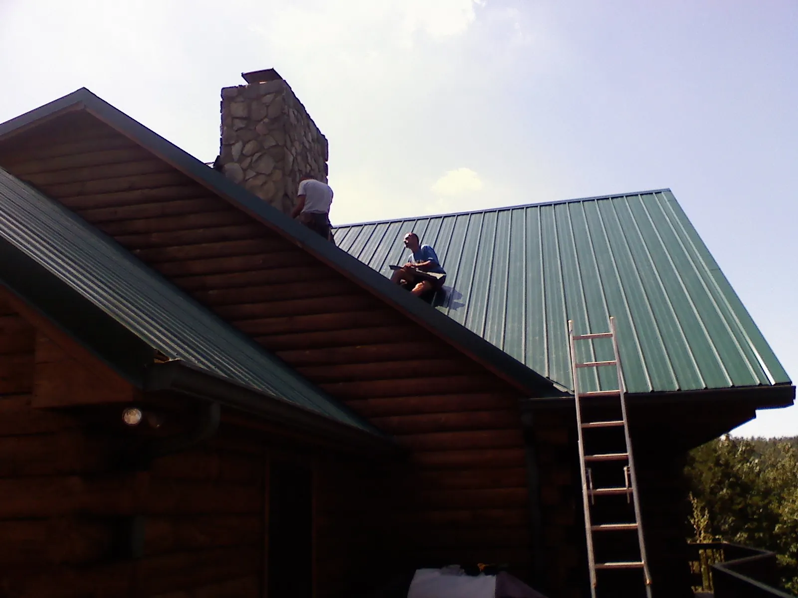 Roofers installing green metal panels beside a stone chimney on a log home in Tivoli